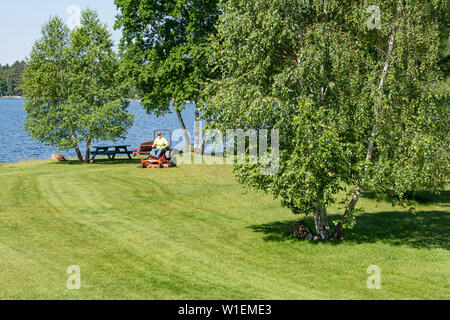 A man cutting grass with a commercial lawn mower at the village park in Speculator, NY USA, with Lake Pleasant in the background. Stock Photo