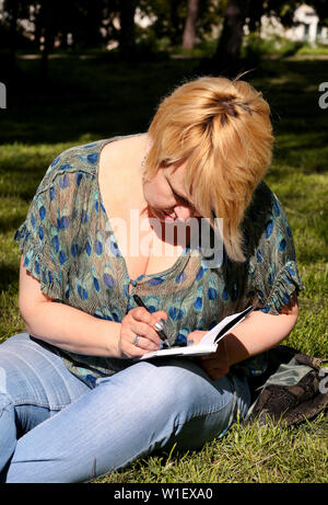 Woman and student sitting on grass, takes notes in notebook, learning and writes thoughts, writes book, she is preparing his final doing homework. Stock Photo