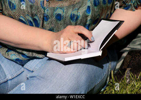 Woman and student sitting on grass, takes notes in notebook, learning and writes thoughts, writes book, she is preparing his final doing homework. Stock Photo