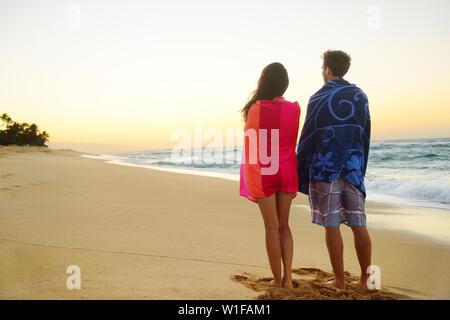 Couple standing in beach sand, towels over shoulders enjoying ocean sunset during travel holidays getaway vacation. Mixed race couple, asian woman, caucasian man. Stock Photo