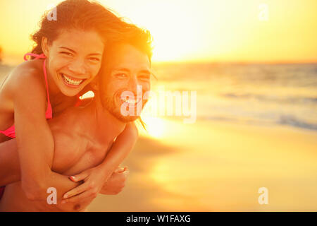 Lovers couple in love having fun dating on beach portrait. Beautiful healthy young adults girlfriend piggybacking on boyfriend hugging happy. Multiracial dating or healthy relationship concept. Stock Photo