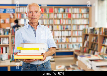 Elderly man holding stack  of books near bookshelves in library interior Stock Photo