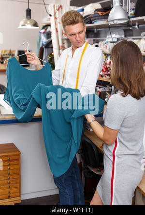 Confident male tailor demonstrating client her order – dress at sewing atelier Stock Photo