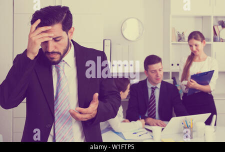 Unhappy and tired man standing at office on background with working colleagues Stock Photo