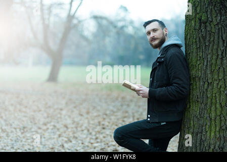 A smiling handsome young hipster man leaning against a tree and reading a book in the park. Stock Photo