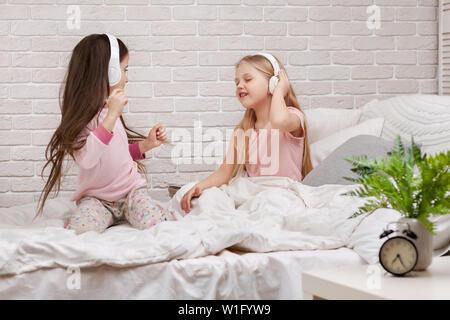 little children girls listening to the music with the headphones and dancing on bed. pajama party and friendship. Stock Photo