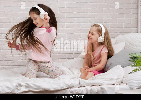 little children girls listening to the music with the headphones and dancing on bed. pajama party and friendship. Stock Photo