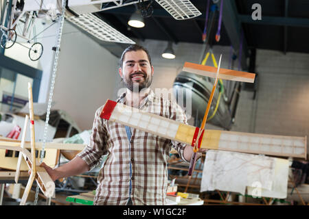 Smiling young bearded man having fun with light plane models in aircraft hangar Stock Photo