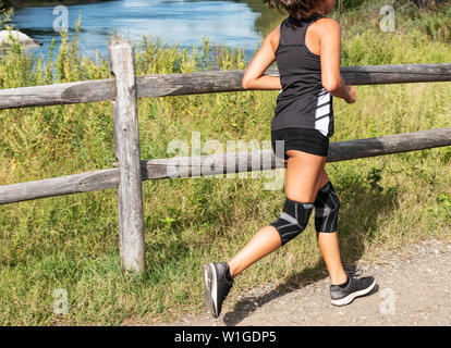 A high school cross country runner is running on a dirt path next two a stream of water a knee brace on both knees. Stock Photo