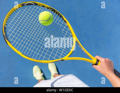 Tennis player holding yellow ball on racket grid. Sports female athlete taking a feet selfie showing running shoes on blue hard court. POV closeup of equipment, neon yellow fashion footwear. Stock Photo