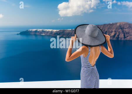 Europe summer vacation travel destination luxury living woman looking at view of Mediterranean Sea and Santorini island Oia village. Elegant tourist lady in fashion back dress and floppy sun hat. Stock Photo