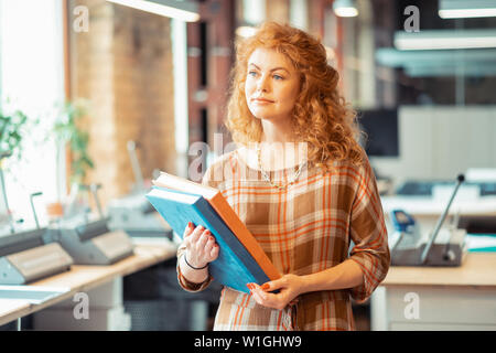 Blue-eyed woman. Blue-eyed curly woman wearing stylish necklace standing in office holding books Stock Photo