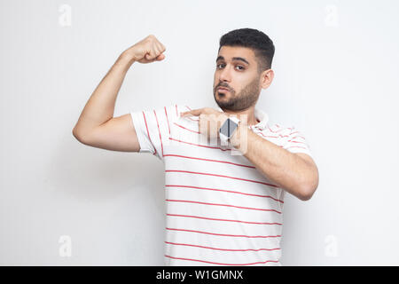 I am strong. Portrait of proud handsome bearded young man in striped t-shirt standing, pointing at his bicep and looking at camera with satisfied face Stock Photo