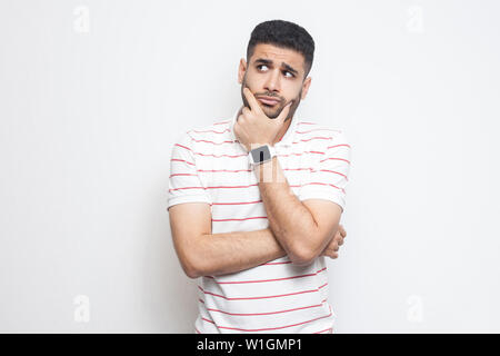 Portrait of pensive handsome bearded young man in striped t-shirt standing, touching his face, looking aside away and thinking about something. indoor Stock Photo