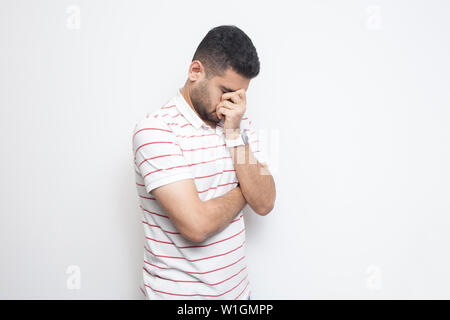 Portrait of sad alone handsome bearded young man in striped t-shirt standing, holding his head down and crying. indoor studio shot, isolated on white Stock Photo