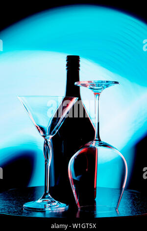 A photo of a wine bottle and two glasses against a light blue stain on a black wall Stock Photo