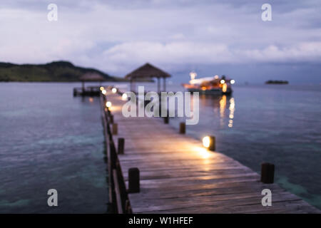 Blurred defocused background of tropical island, pier and yacht in twilight with evening lights in bokeh. Marine travel and vacations concept. Stock Photo