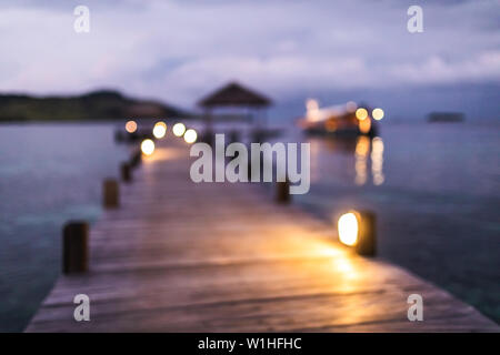 Blurred defocused background of tropical island, pier and yacht in twilight with evening lights in bokeh. Marine travel and vacations concept. Stock Photo