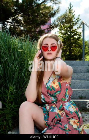 Young attractive woman. Girl in the red sunglasses and color dress. Young woman sits on the steps in the summer city park. The blue sky and urban back Stock Photo