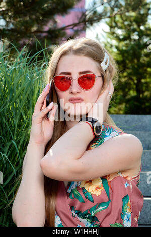 Young attractive woman. Girl in the red sunglasses and color dress. Young woman sits on the steps in the summer city park. The blue sky and urban back Stock Photo