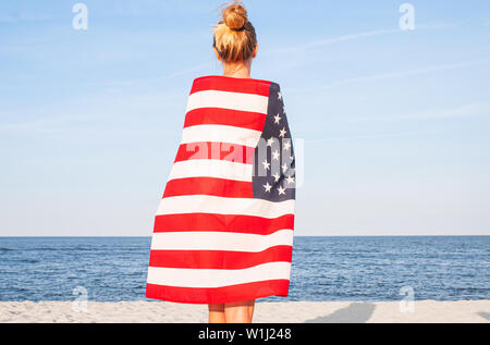 Beautiful patriotic woman with American flag on the beach.  USA Independence day, 4th July. Freedom concept, view from the back.