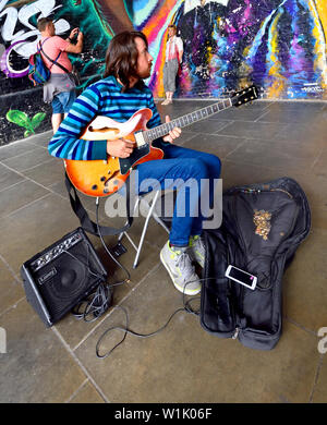 London, England, UK. Busker playing the guitar in front of a mural in Southwark - people taking a photo in the background Stock Photo