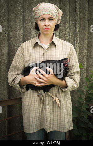 woman farmer in men's clothing, holding a black chicken in her hands in the rural scene. Stock Photo