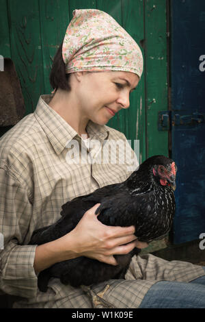 woman farmer in men's clothing, holding a black chicken in her hands in the rural scene. Stock Photo