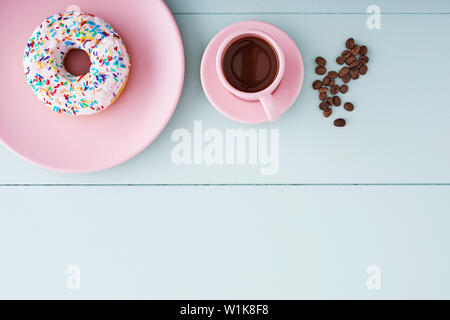 One white donut or doughnut in a pink plate, cappuccino in a pink coffee cup and coffee beans on a pastel blue wooden table with copy space. Top view. Stock Photo