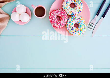 Colorful donuts in a pink plate with coffee cup, fork, knife and marshmallows on a pastel blue wooden table with copy space. Top view. Stock Photo