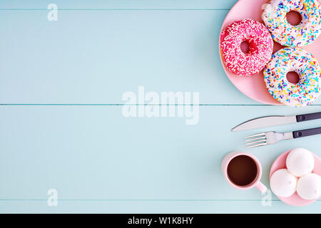 Colorful donuts in a pink plate with coffee cup, fork, knife and marshmallows on a pastel blue wooden table with copy space. Top view. Stock Photo