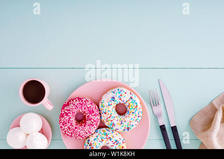 Colorful donuts in a pink plate with coffee cup, fork, knife and marshmallows on a pastel blue wooden table with copy space. Top view. Stock Photo