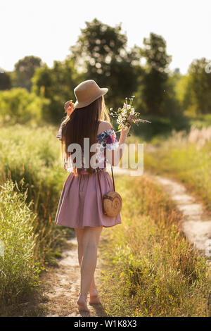 a slender girl in a straw hat and with a straw handbag turned away and holding a bouquet of daisies in the meadow. Summer sunset, the concept of happi Stock Photo