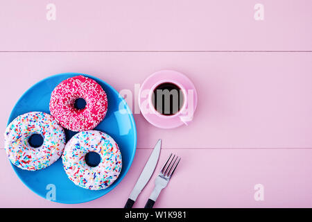 Colorful donuts in a turquoise plate with pink coffee cup, fork and knife on a pastel pink wooden table background with copy space. Overhead view. Stock Photo