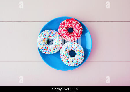 Three colorful donuts or doughnut in a turquoise plate on pastel pink wooden table background. Overhead view. Stock Photo