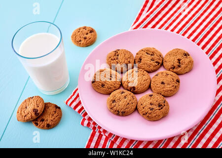 A glass full of milk, chocolate chip cookies in a pink plate and a red tablecloth on blue wooden table. Close up view from top. Stock Photo