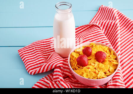 Bowl of corn flakes cereal with strawberry topping, a bottle of milk and tablecloth on blue wooden country style table. View from above. Stock Photo