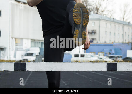 A man in black clothes is exercising outdoors with a barrier. fitness athlete on the sports field. training with hurdle. warm up stretching legs. body Stock Photo