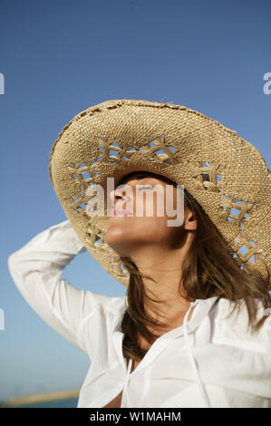 Young woman with straw hut standing on the beach, eyes closed, side view Stock Photo
