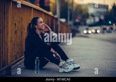 Young woman in black sports outfit listening to music and resting after running on the bridge in the city, at night