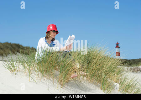 Woman wearing a red hut sitting at beach while reading a book, List-Ost lighthouse in background, Ellenbogen, List, Sylt, Schleswig-Holstein, Germany Stock Photo