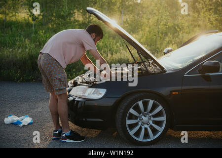 The guy in the pink t-shirt checks the engine of a black car lifting the hood in the country outside the road Stock Photo