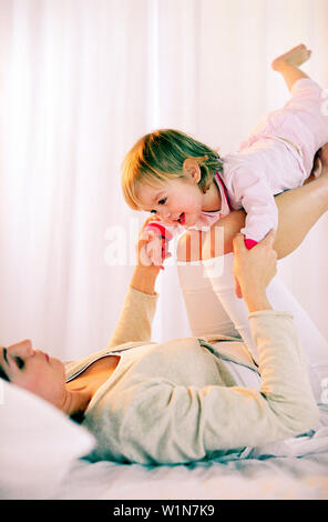 Toddler girl balancing on mother's knees on bed, side view Stock Photo