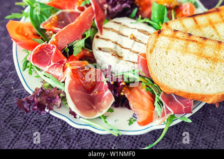 Tasty mixed salad dish with grilled camembert cheese, prosciutto ham, organic tomato and fresh green leaves. Healthy dilicious meal. Isolated on black Stock Photo