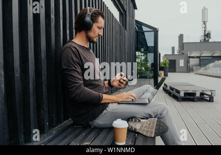 Photographer freelancer siting i the city wih hot tea or coffe listens to music on headphones, backpac and works on a laptop Stock Photo