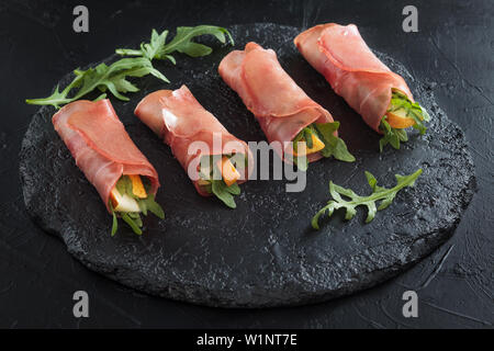 Prosciutto rolls stuffed with arugula and pears on black slate. Stock Photo