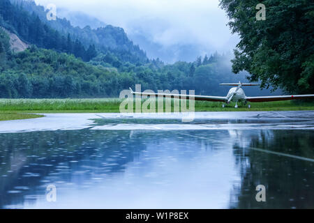 With tarpaulin-covered sports aircraft is located in power rain on the tarmac of the gliding school in unterwössen, forest and mountains in the backgr Stock Photo