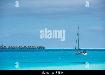 A small anchored sailboat sits in turquoise water in the foreground, while in the background a line of thatch-roofed overwater bungalows on stilts fro Stock Photo
