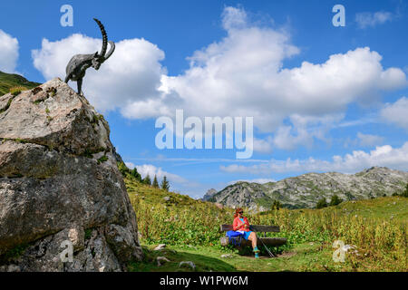 Woman while hiking sits on bank, Capricorn sculpture stands next to it, Lech source rocks, lechweg mountains, Vorarlberg, Austria Stock Photo