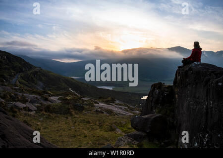 A woman sits relaxed on a rock and admires the sunset over the mountain pass with a vast panoramic landscape, seen from while walking the Dingle Way, Stock Photo